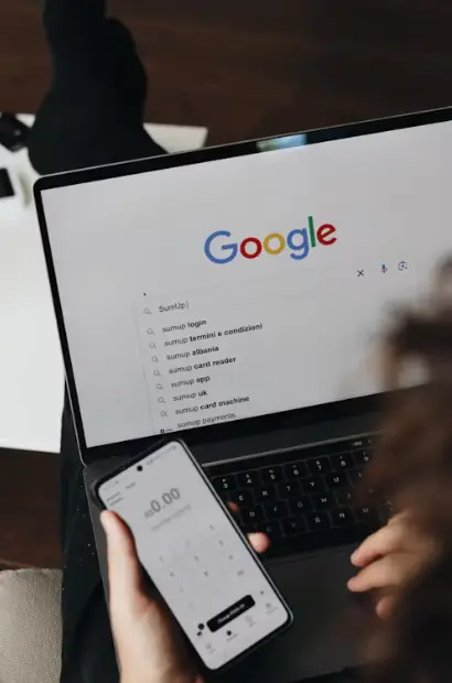 a woman holding a mobile phone displaying search results and looking at computer monitor of Google Search
